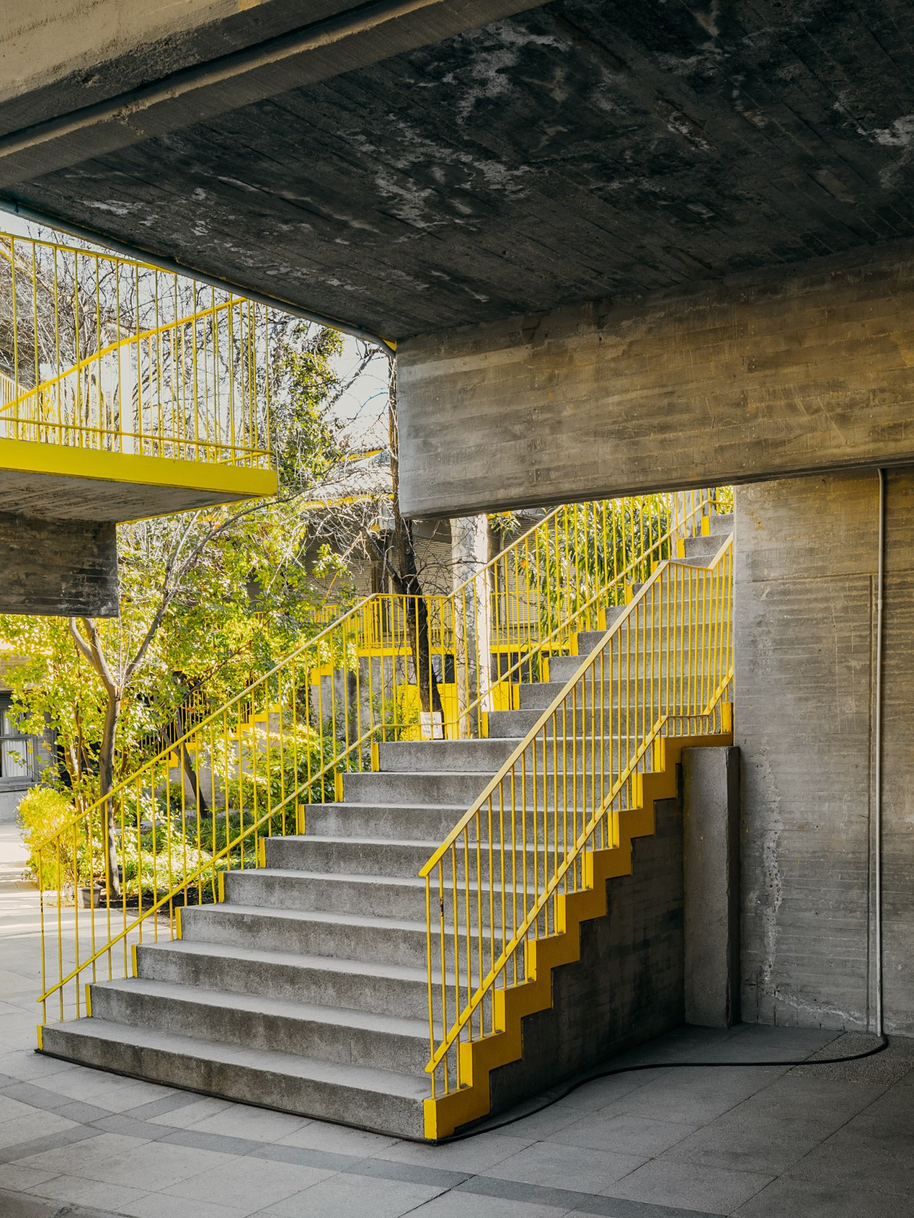 Brutalist concrete staircase with bright yellow metal railings and steps, surrounded by dark concrete beams and green foliage.