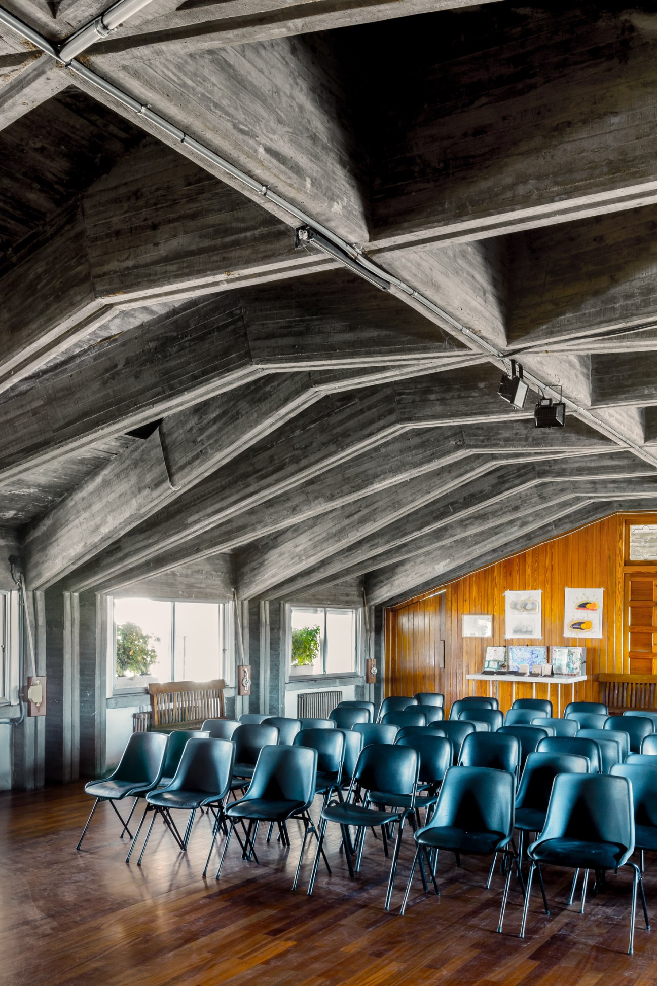 School theatre interior with blue seating beneath brutalist concrete beams and wooden wall paneling.