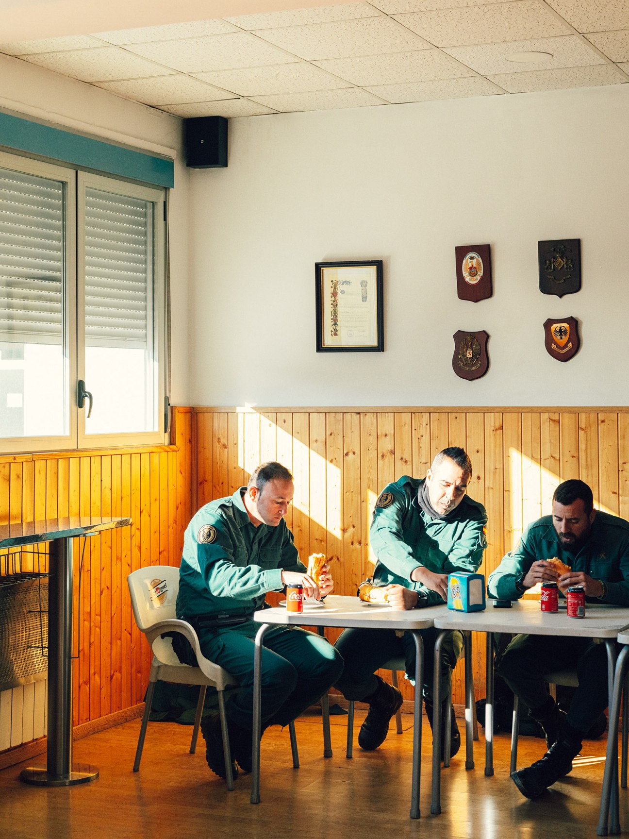 Three Guardia Civil agents in green uniforms eat at a table in a wooden-paneled cafeteria during a break.