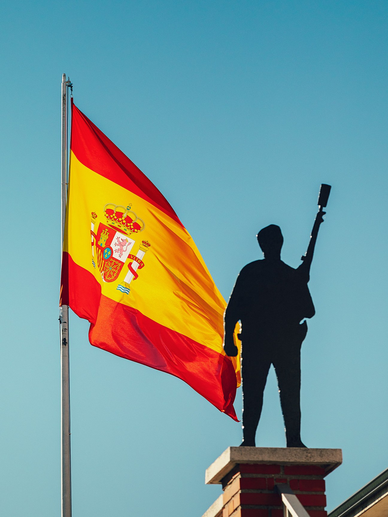 Spanish flag flying next to silhouette statue of armed figure on Guardia Civil base.