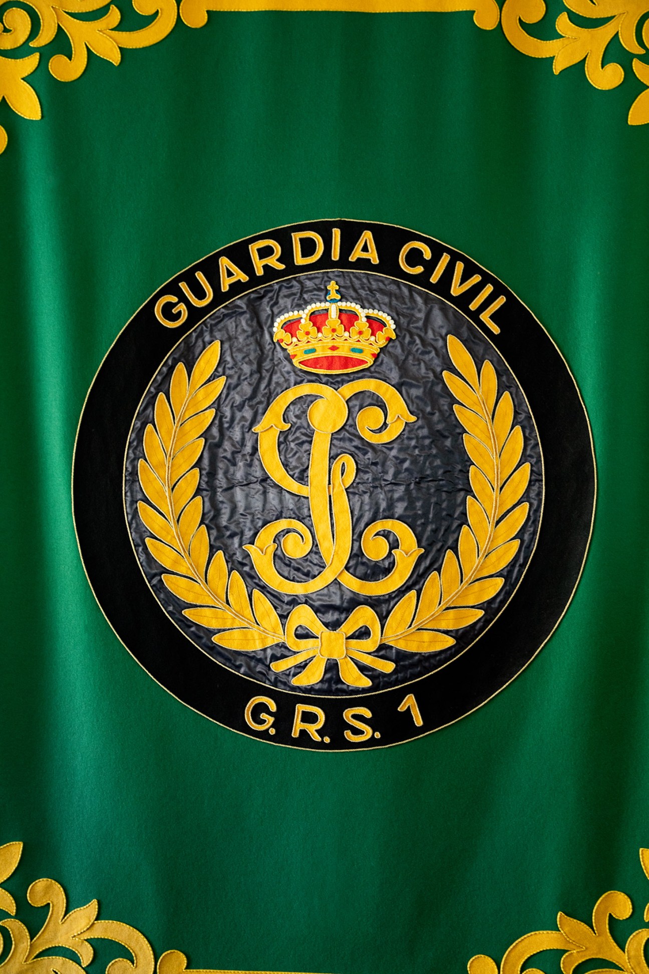 GRS emblem featuring gold laurel wreaths, a crown, and ornate scrollwork on a green and black circular seal.