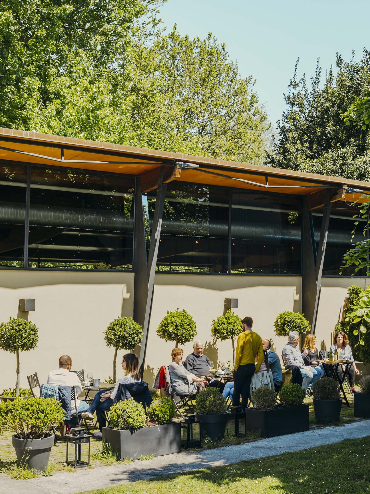 Outdoor terrace with diners seated under yellow umbrellas and shade structures surrounded by topiary plants.
