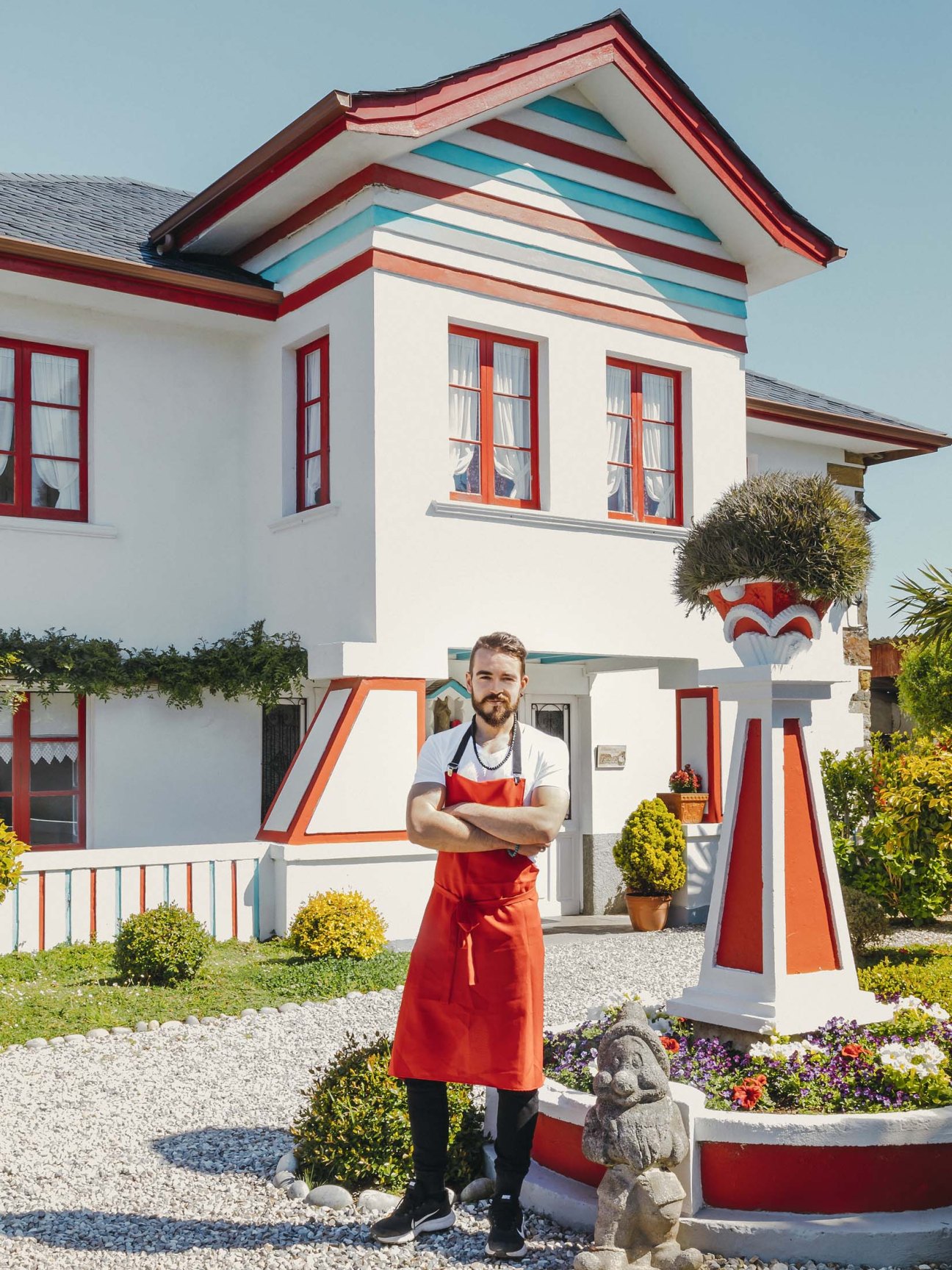 Pastry chef Jhonatan González Ovalle stands in a red apron outside his colorful cottage bakery with flower beds and topiary.