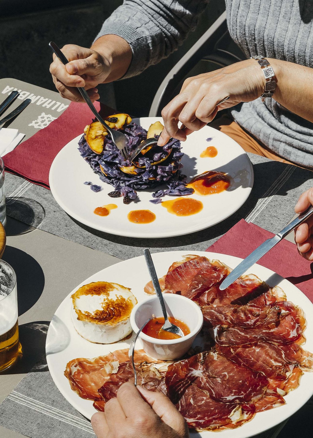 Hands with forks over plates of cured meat and purple cabbage salad on outdoor dining table