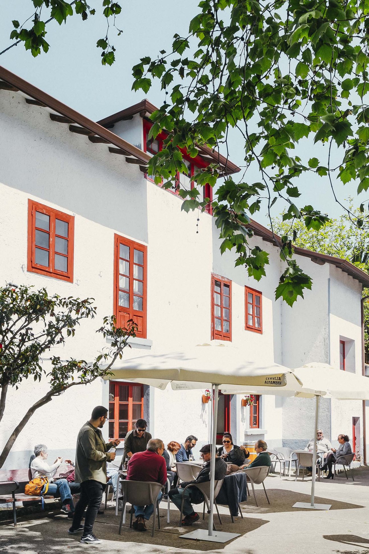 Diners seated at outdoor tables under a cream umbrella in front of a white building with red trim and shutters covered in climbing ivy.