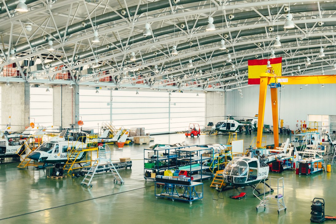 Guardia Civil helicopter repair hangar with multiple aircraft undergoing maintenance beneath industrial ceiling and Spanish flag.