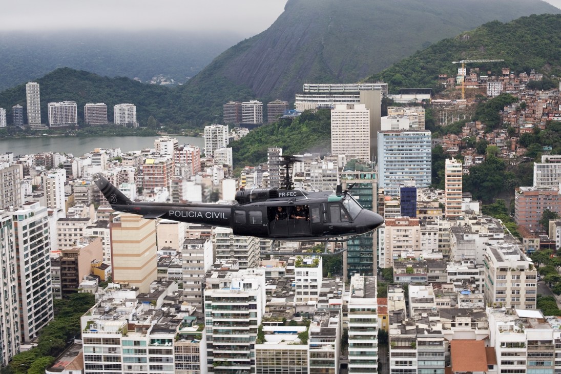 A black Policia Civil UH-1H helicopter flies over Rio de Janeiro's Copacabana district with mountains and favelas visible below.