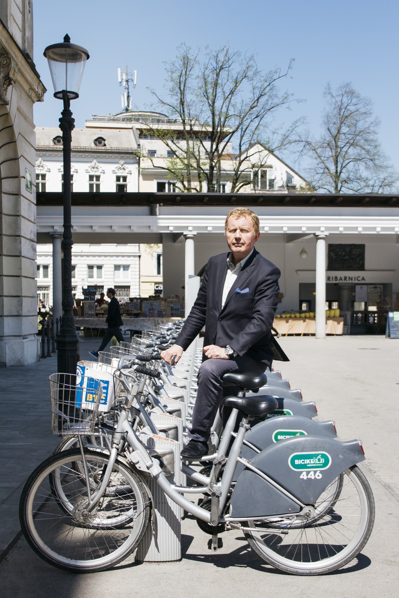 Man in dark blazer sits on a bike-sharing bicycle in an urban plaza with white neoclassical buildings in background.
