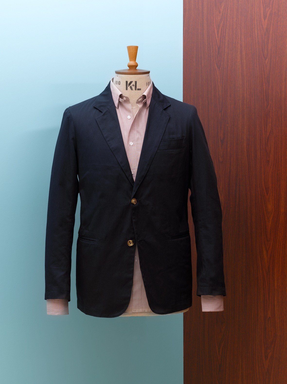 Navy blazer with pink shirt on tailoring mannequin against blue and wood-grain background.