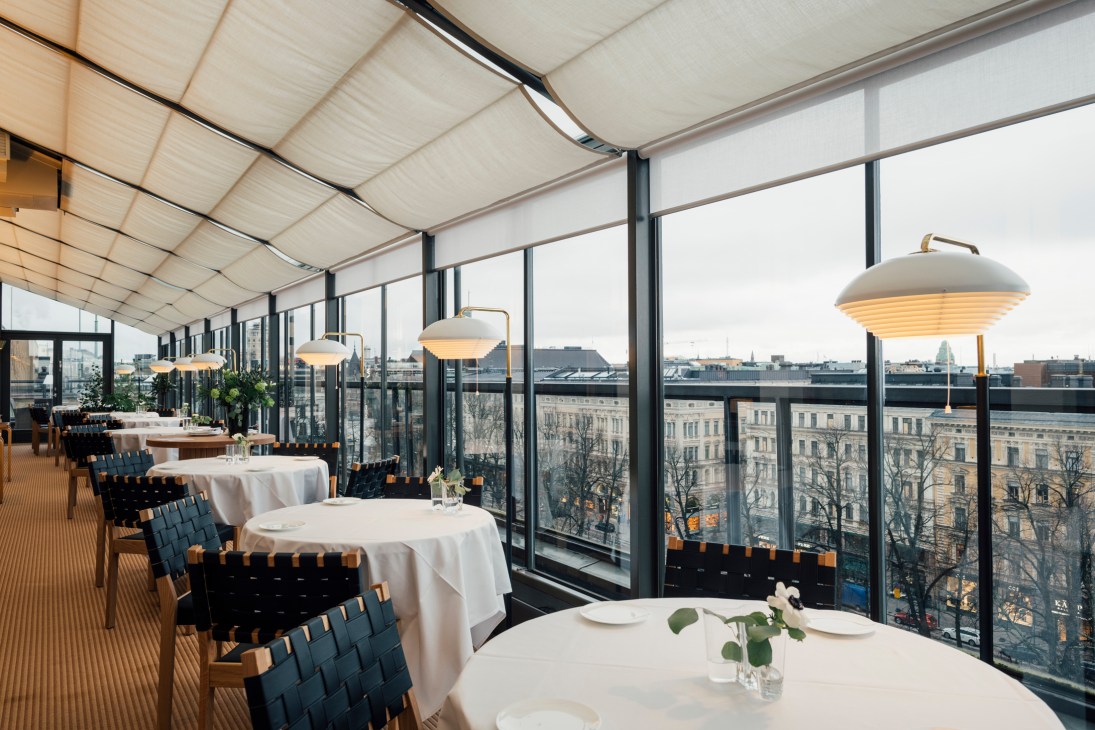Modern restaurant dining room with floor-to-ceiling windows, round tables with white tablecloths, and pendant lamps