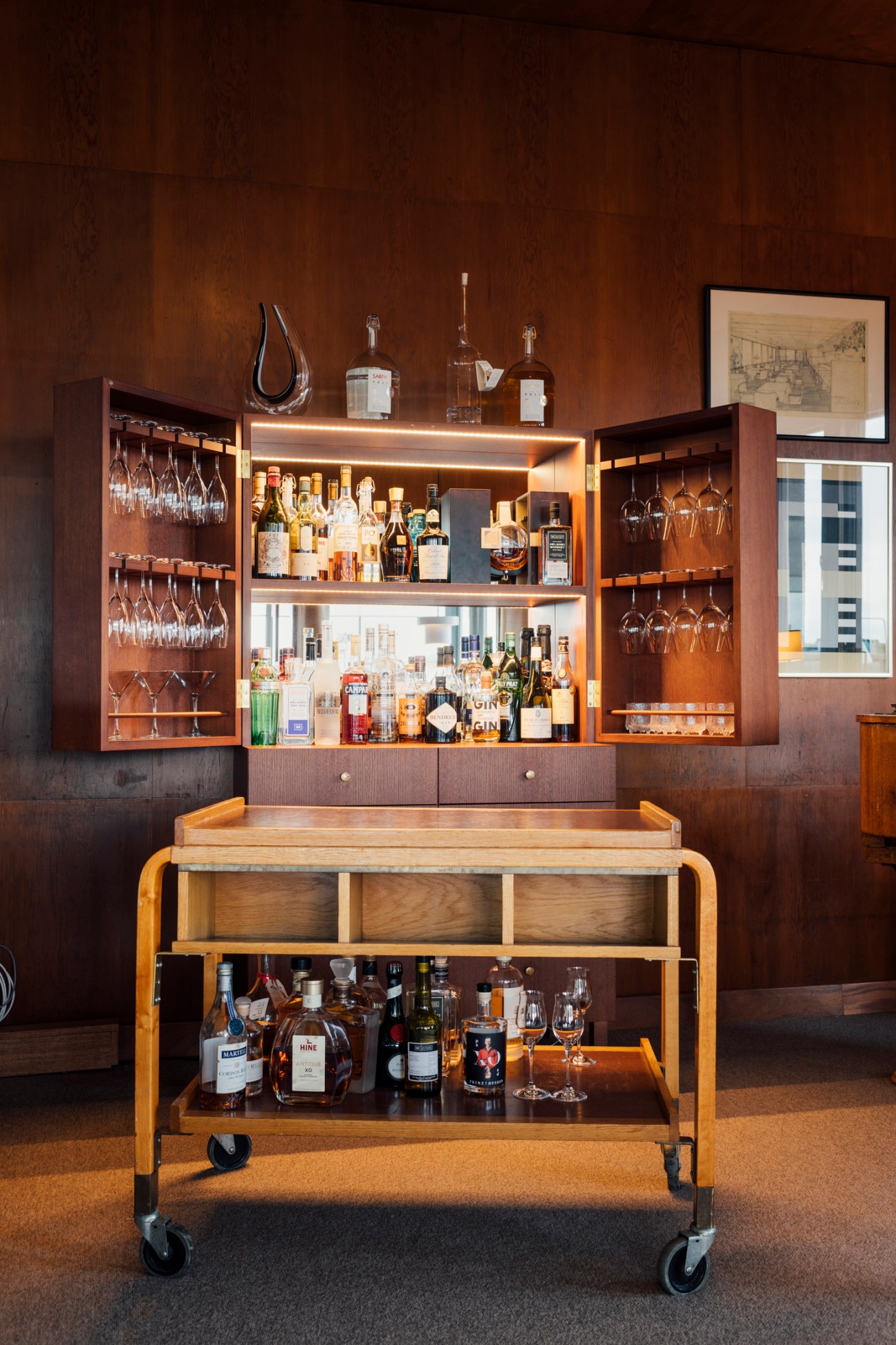 Elegant wooden bar cart with bottles of spirits and glassware positioned in front of built-in bar cabinet