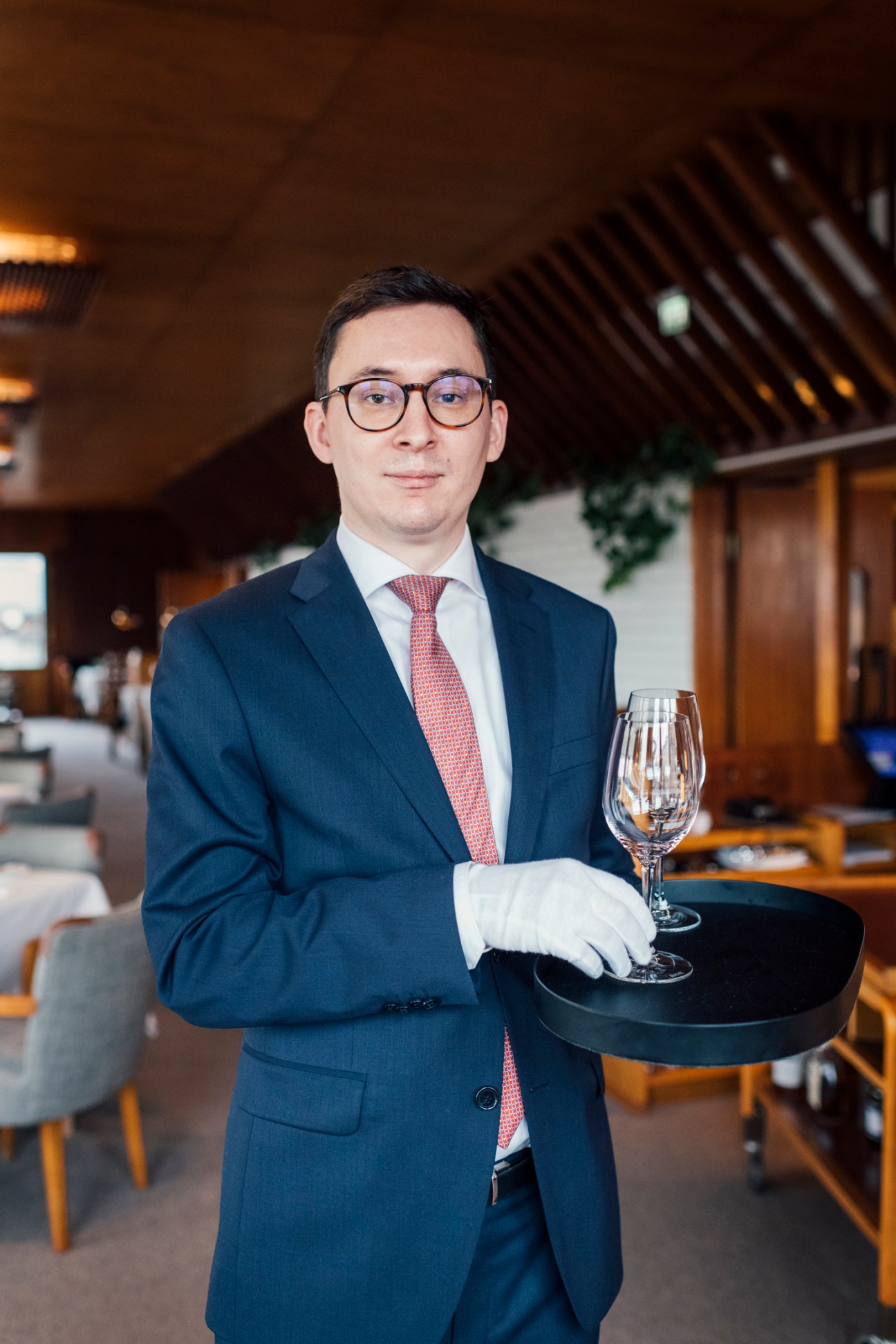 Waiter Anssi Bourgoin in dark suit and glasses holding wine glass on tray in restaurant dining room