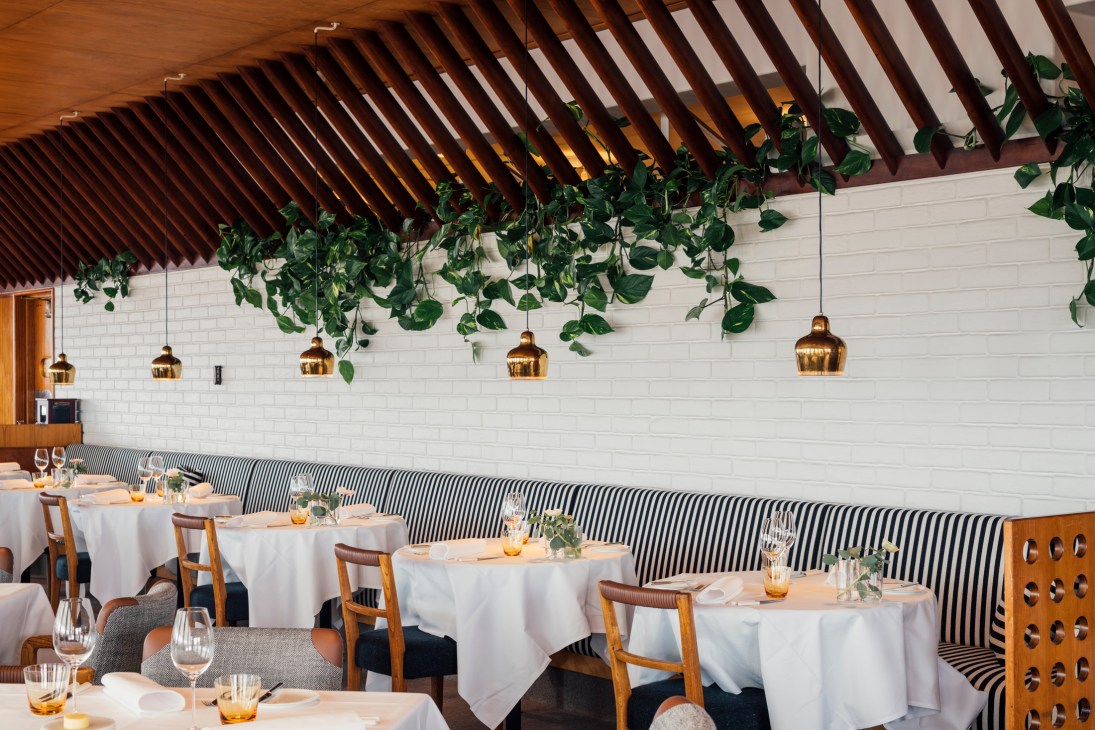 Restaurant dining room with wooden ceiling, striped banquette seating, white tablecloths, and hanging brass pendant lights