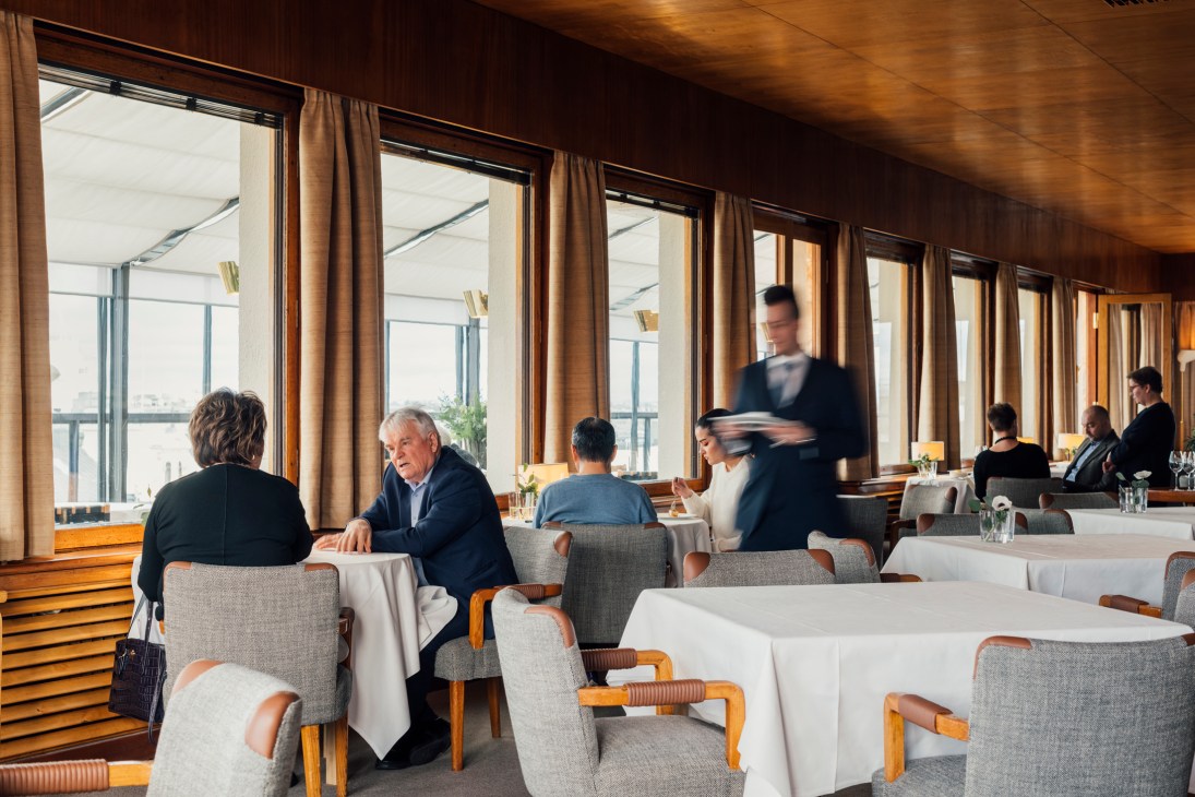 Diners seated at white-clothed tables with gray upholstered chairs in a wood-paneled restaurant with large windows