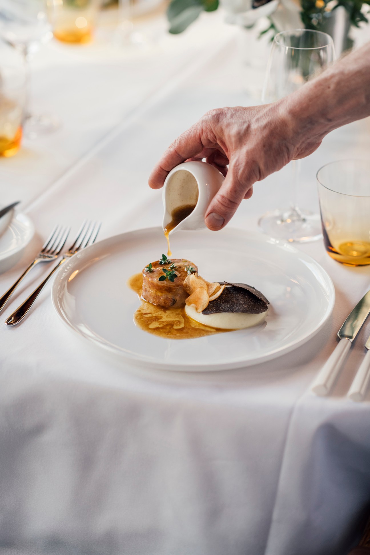 Hand pouring sauce from white ceramic pitcher onto plated guinea fowl ballotine with garnishes
