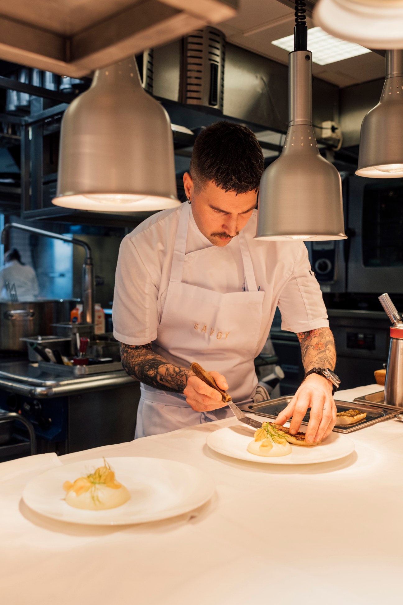 Chef in white uniform with tattooed arms plating food under pendant lights in professional kitchen