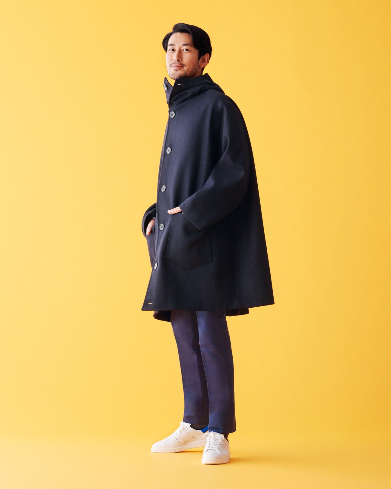 Man wearing a navy coat and trousers with white trainers against a yellow background.