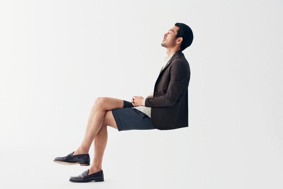 Man in brown jacket, shorts and slip-on shoes sitting in profile against white background.