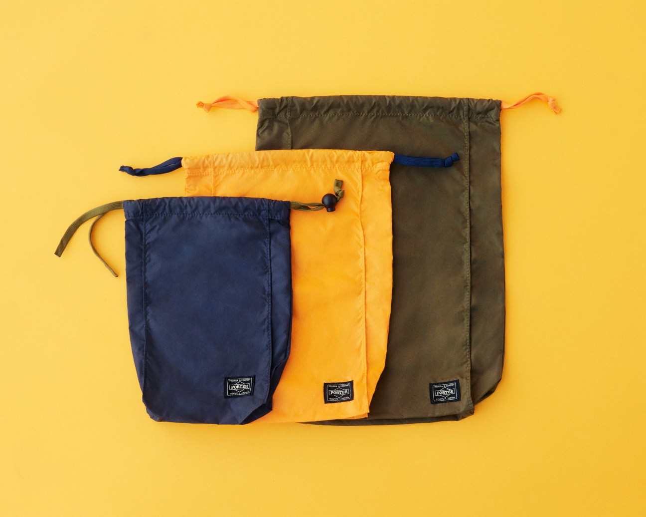 Three Porter nylon drawstring pouches in navy, yellow and olive against a yellow background.