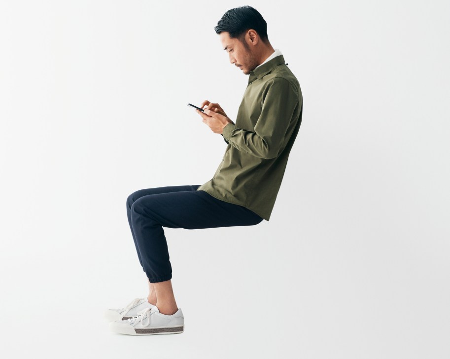 Man in olive jacket and navy trousers looking at smartphone while sitting.
