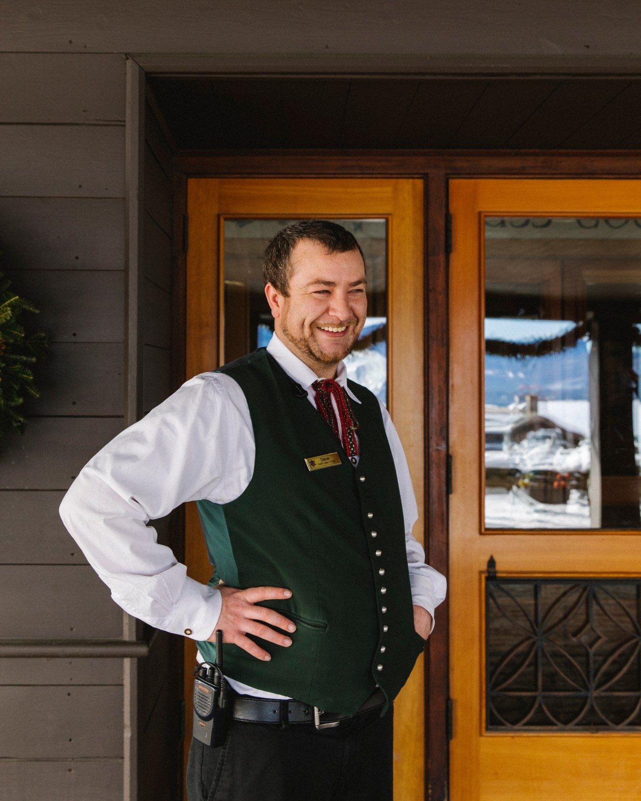 Smiling man in traditional Austrian green vest and white shirt standing in front of wooden lodge entrance