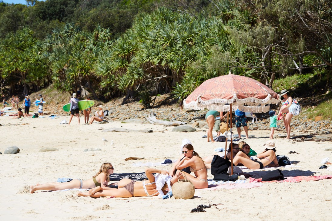 Beachgoers relaxing on sand at Spooky Beach with a pink umbrella and palm trees in background.
