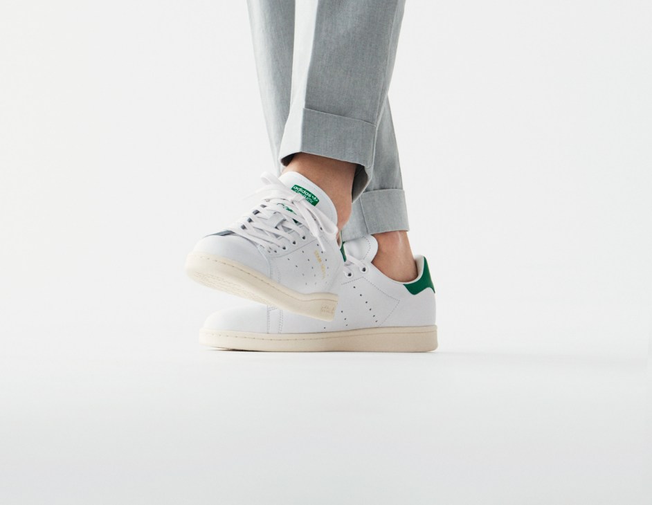 Person wearing grey trousers and white Adidas trainers with green heel tab.