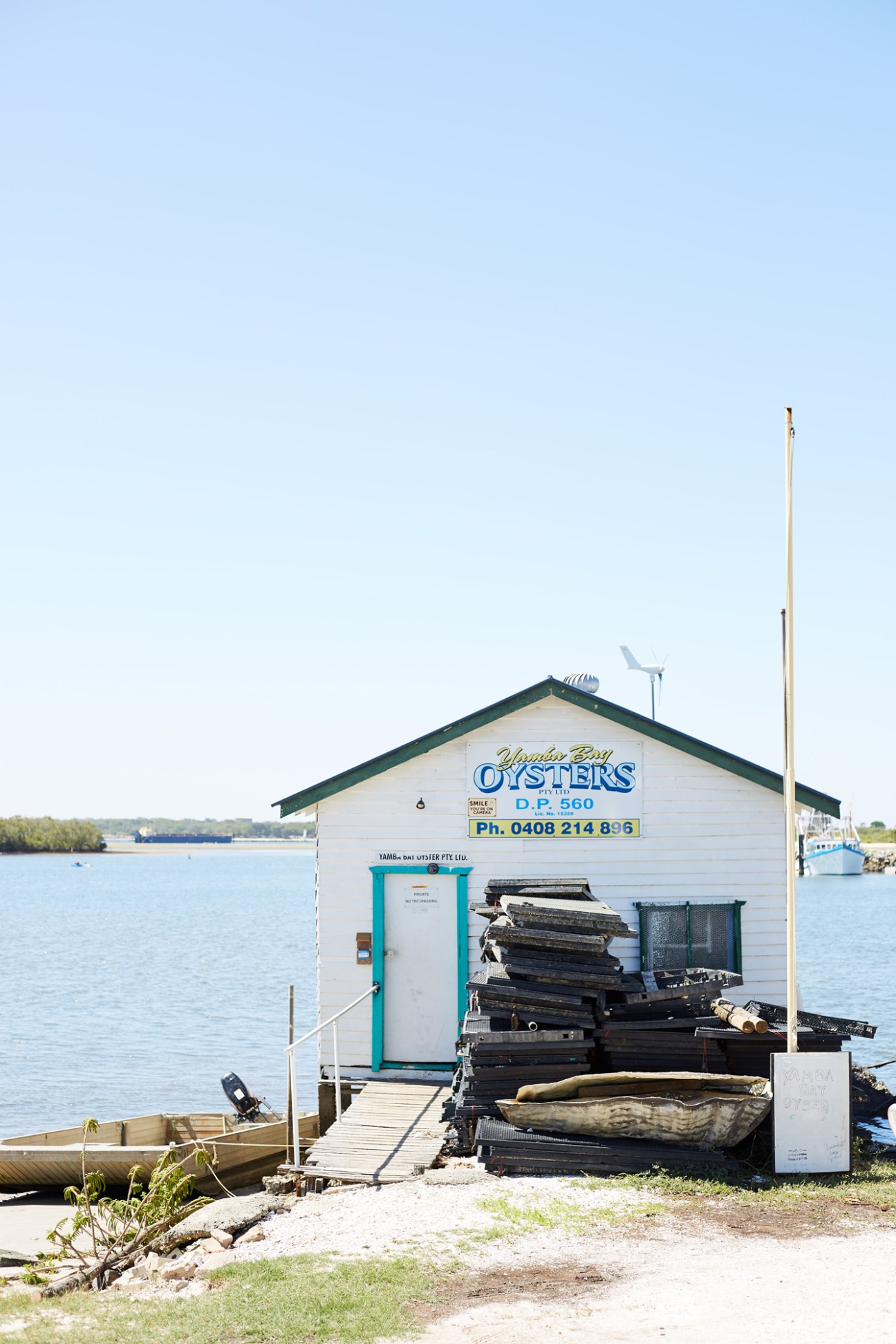 White bayside oyster shack with green trim and signage for Yamba Bay Oysters, surrounded by black oyster traps and wooden dock.