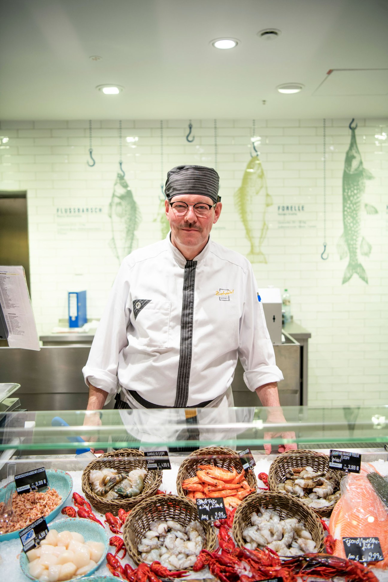 Fish department worker in white chef's coat standing behind counter with seafood displays in woven baskets.