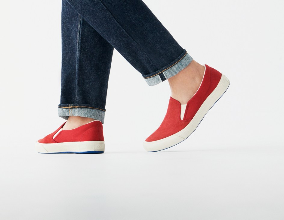 Person wearing dark blue rolled-cuff jeans and red and cream slip-on sneakers by MoonStar against white background.