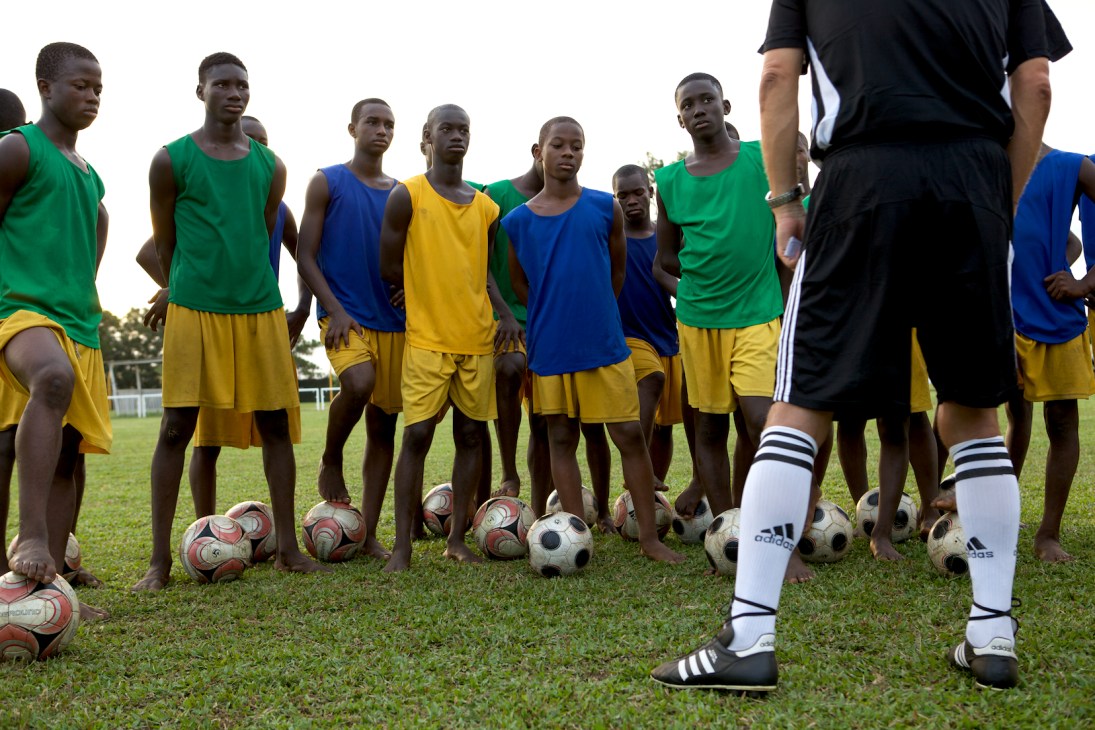 Young football academy students in colored training bibs listen to their coach during a field session.