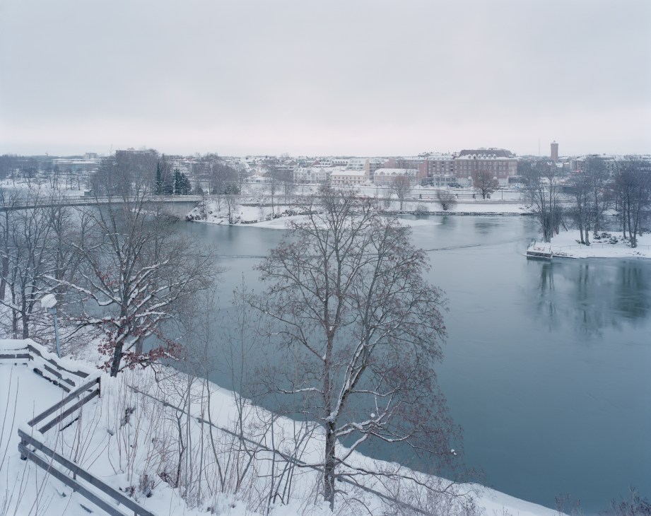 Snowy winter landscape of Trollhättan showing the Göta river, bare trees, and industrial buildings across the water.