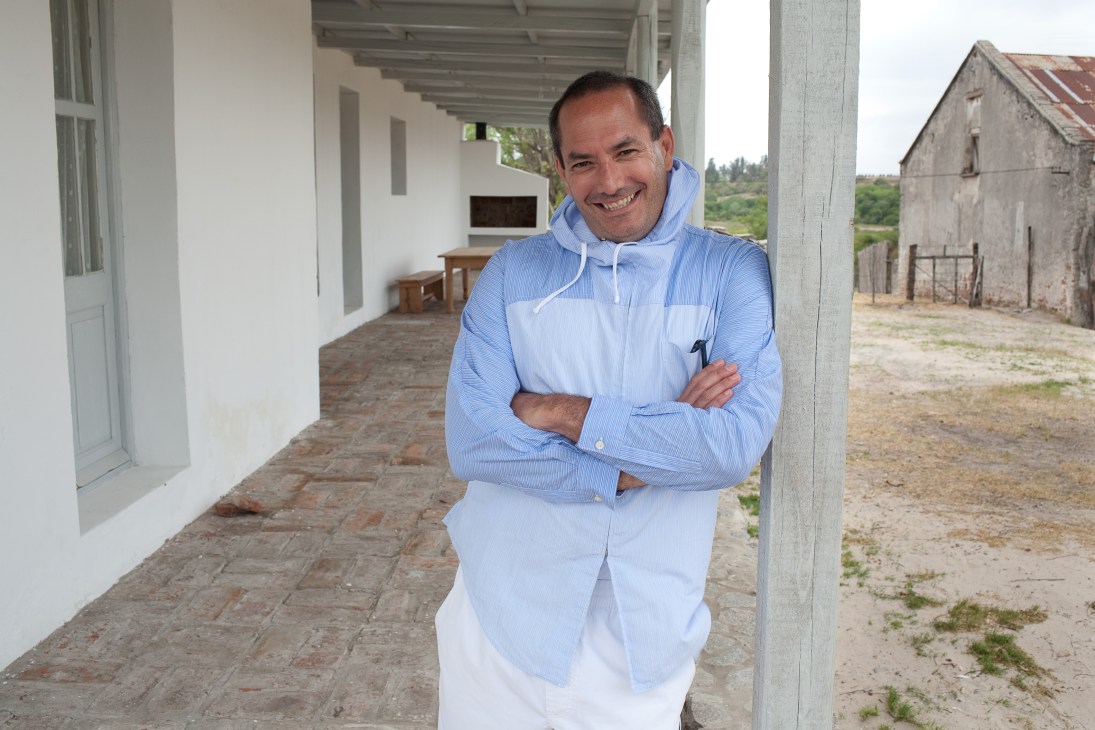 Argentine music producer Tutu Gianakis stands smiling with arms crossed on a covered porch of a rural property in Carmelo, Uruguay.