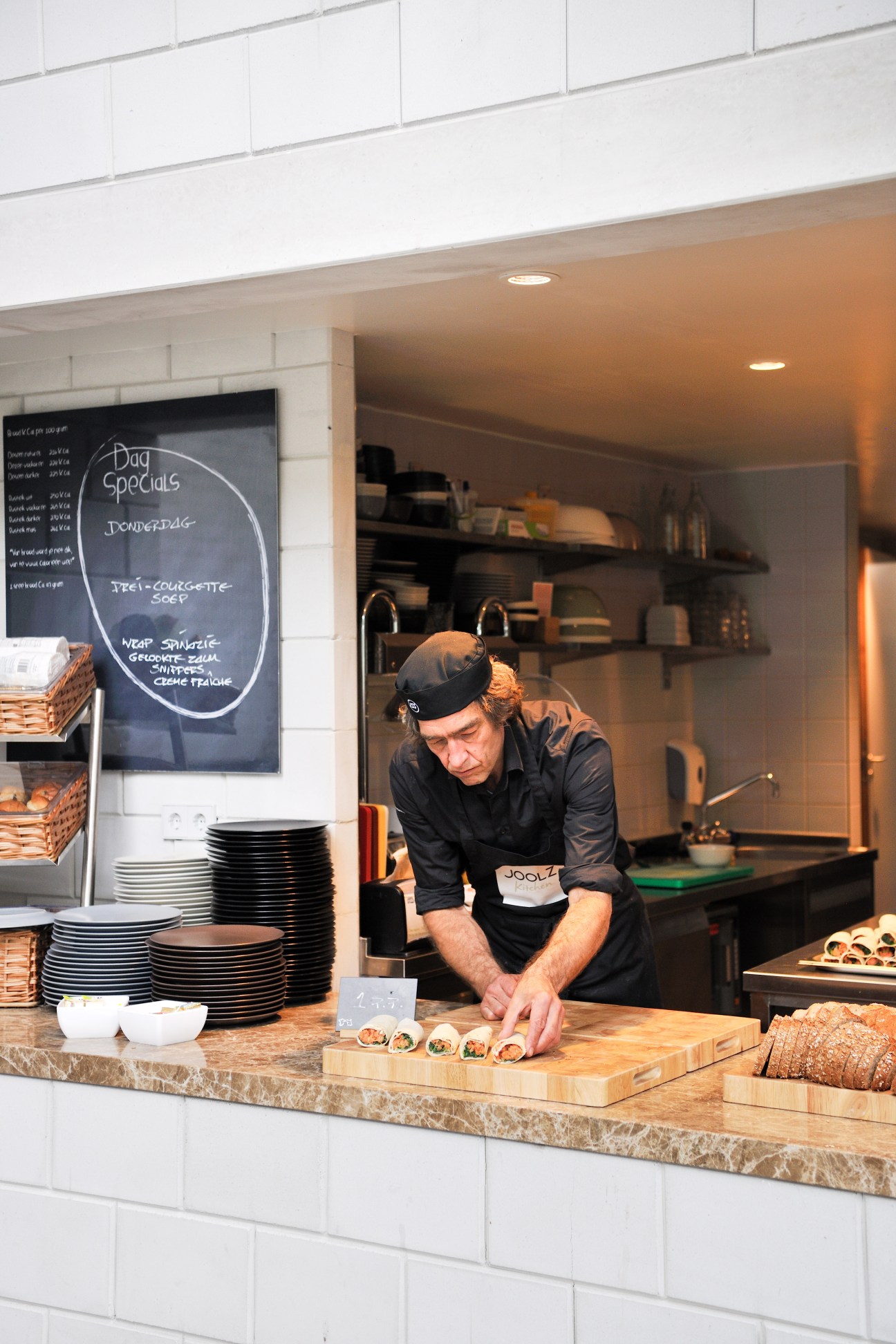 Chef in black uniform and cap preparing food on wooden cutting board in modern open kitchen with white subway tiles