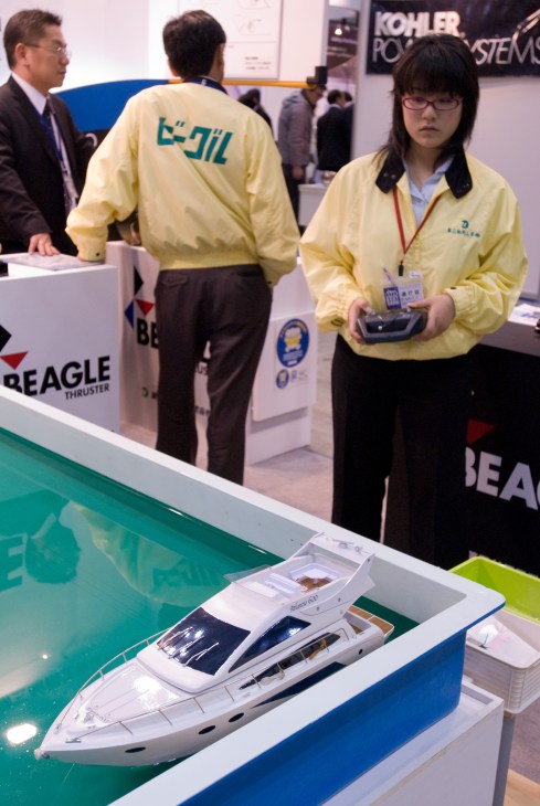 A radio-controlled model boat displayed on a turquoise platform with staff at a boat show booth.