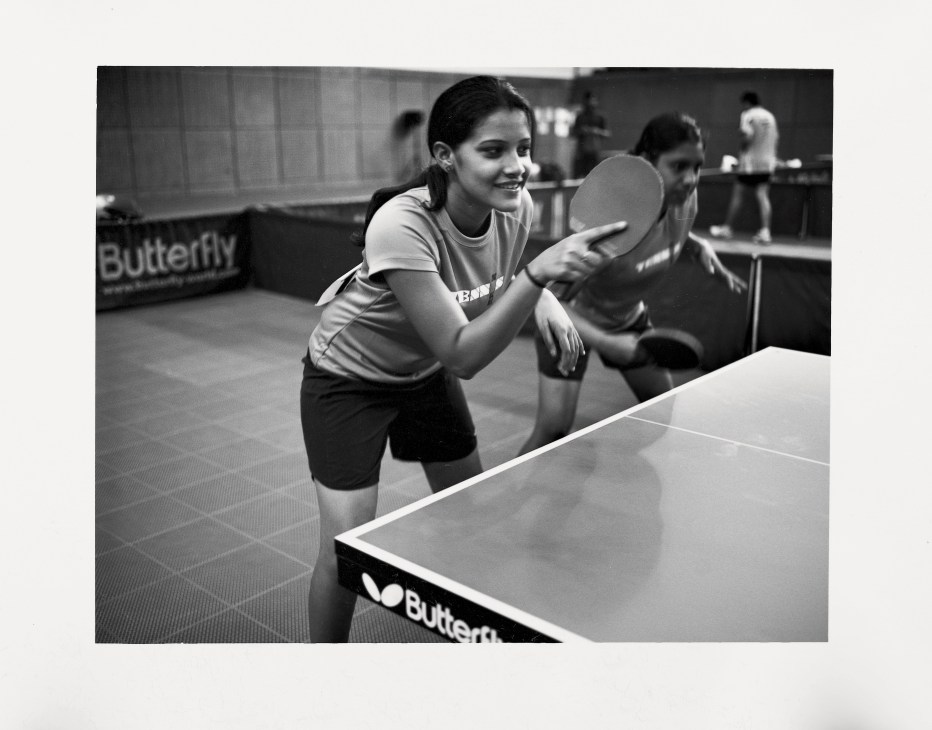 Woman in white shirt playing table tennis, holding paddle mid-rally at indoor facility.