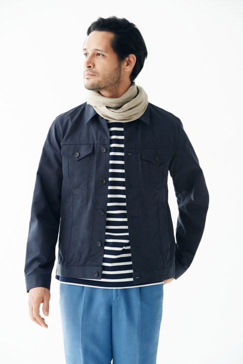 Man wearing navy jacket, striped shirt, blue trousers and beige scarf against white background.