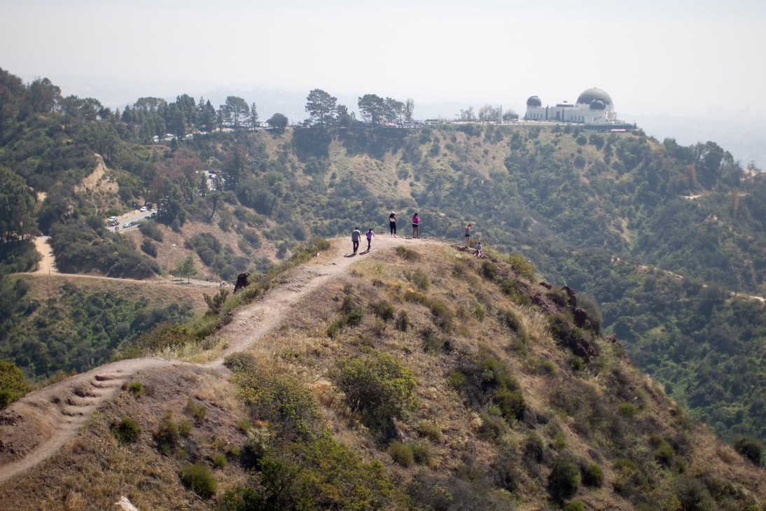 Hikers standing on a mountain ridge overlooking a forested valley with the Griffith Observatory visible in the distance.