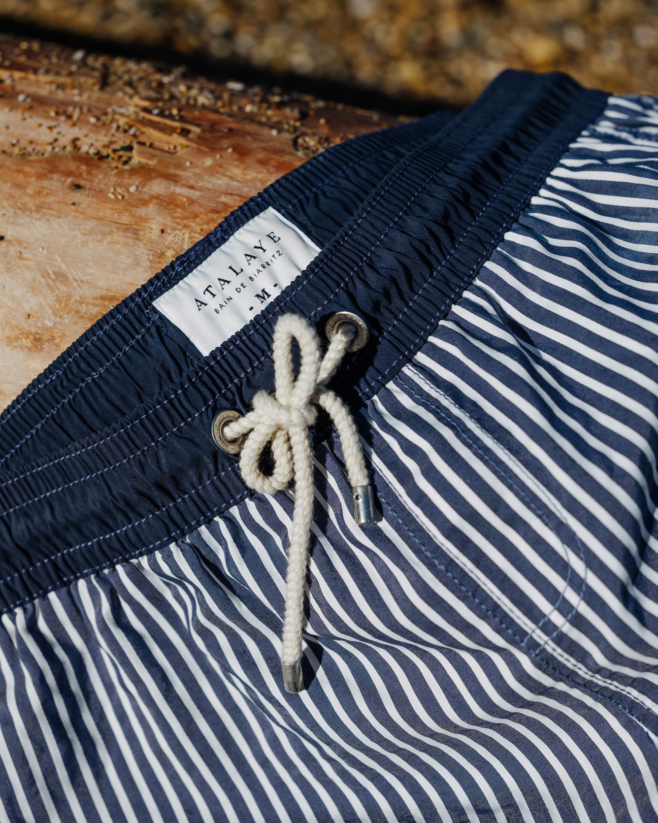 Atalaye swim shorts with navy and white striped pattern and white drawstring on weathered wood.