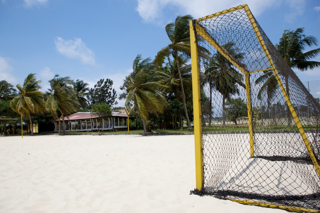Yellow goal frame on a sandy beach football pitch with palm trees and a pavilion in the background.