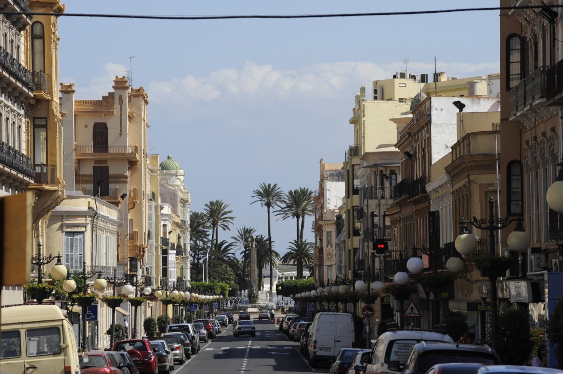 A tree-lined street in Melilla with modernist buildings, parked cars, and palm trees leading toward Plaza España.