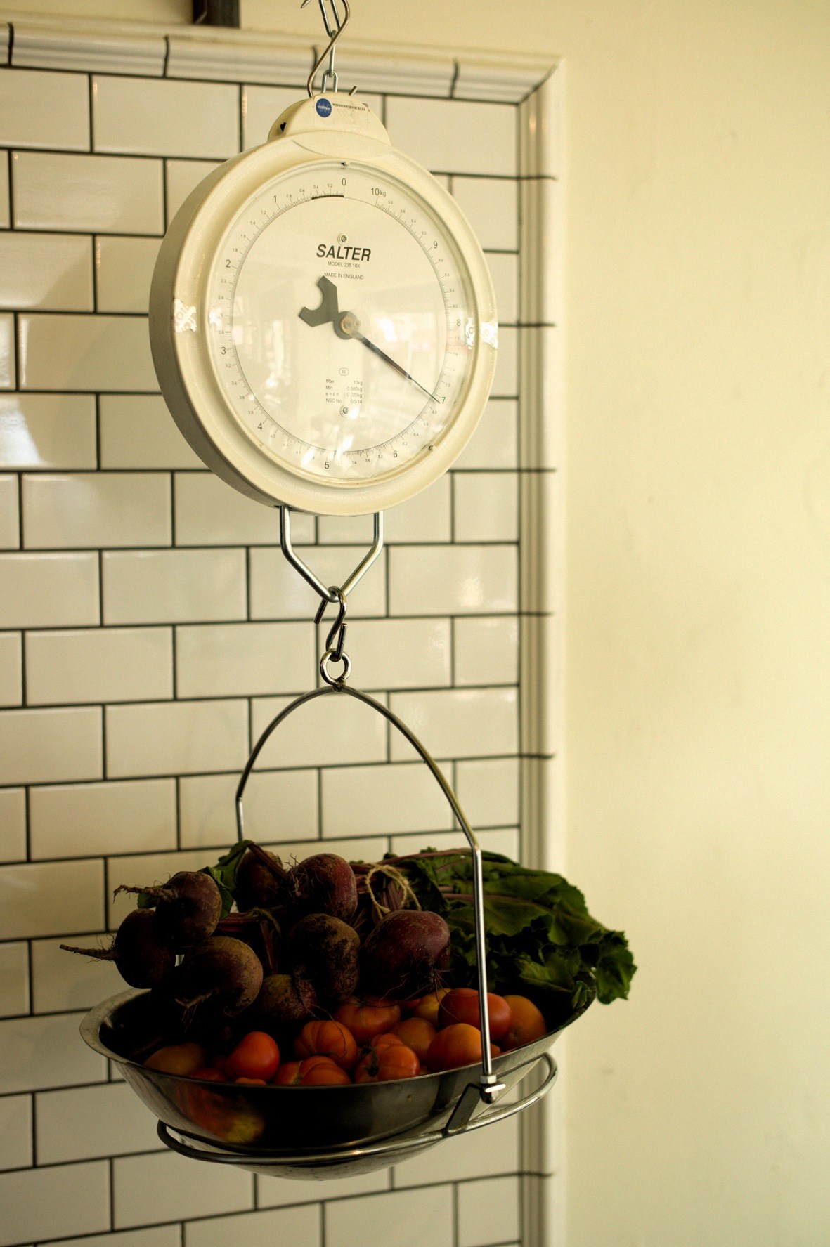 A vintage Salter hanging scale with fresh produce in the basket against subway tile wall.