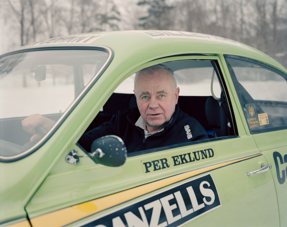 Rally driver Per Eklund sits in a lime green Saab 96 V4 rally car in snowy conditions.