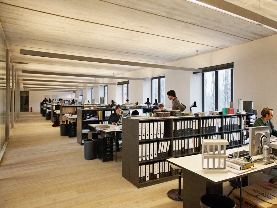 Open-plan design studio with wooden floors, staff at desks, and exposed concrete ceiling beams.