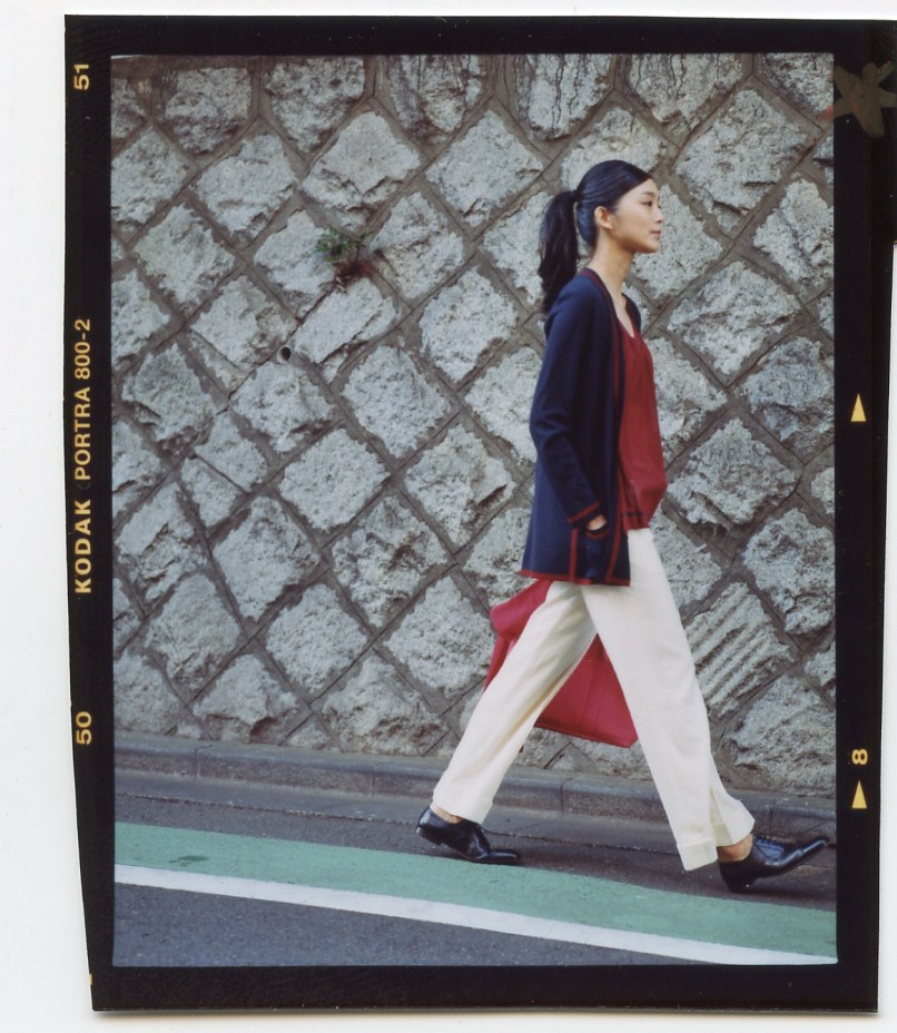 Woman in navy cardigan and white trousers walking past textured stone wall in Tokyo.