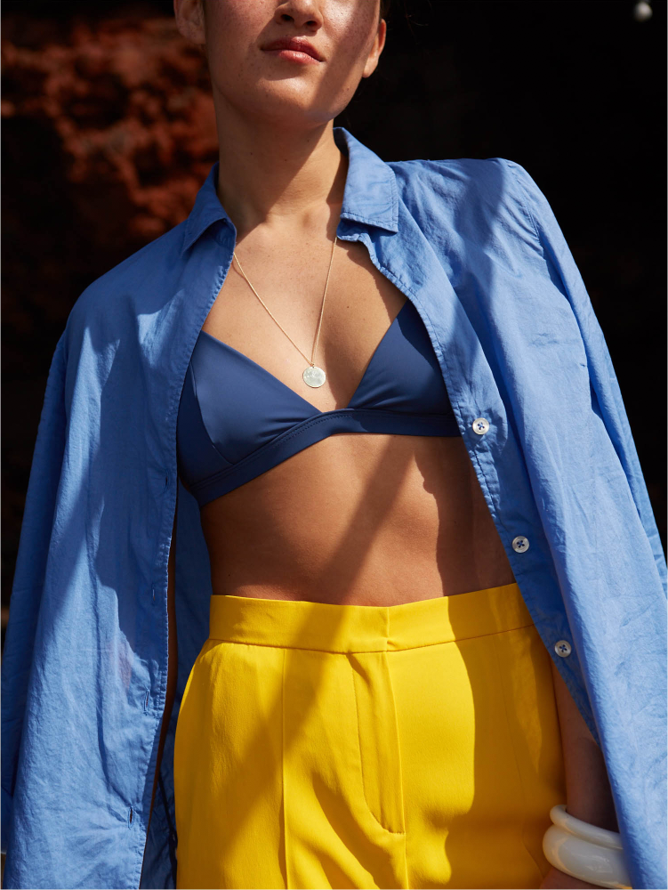 Woman wearing a blue linen shirt, navy bikini top, yellow trousers, and silver necklace and bangle.