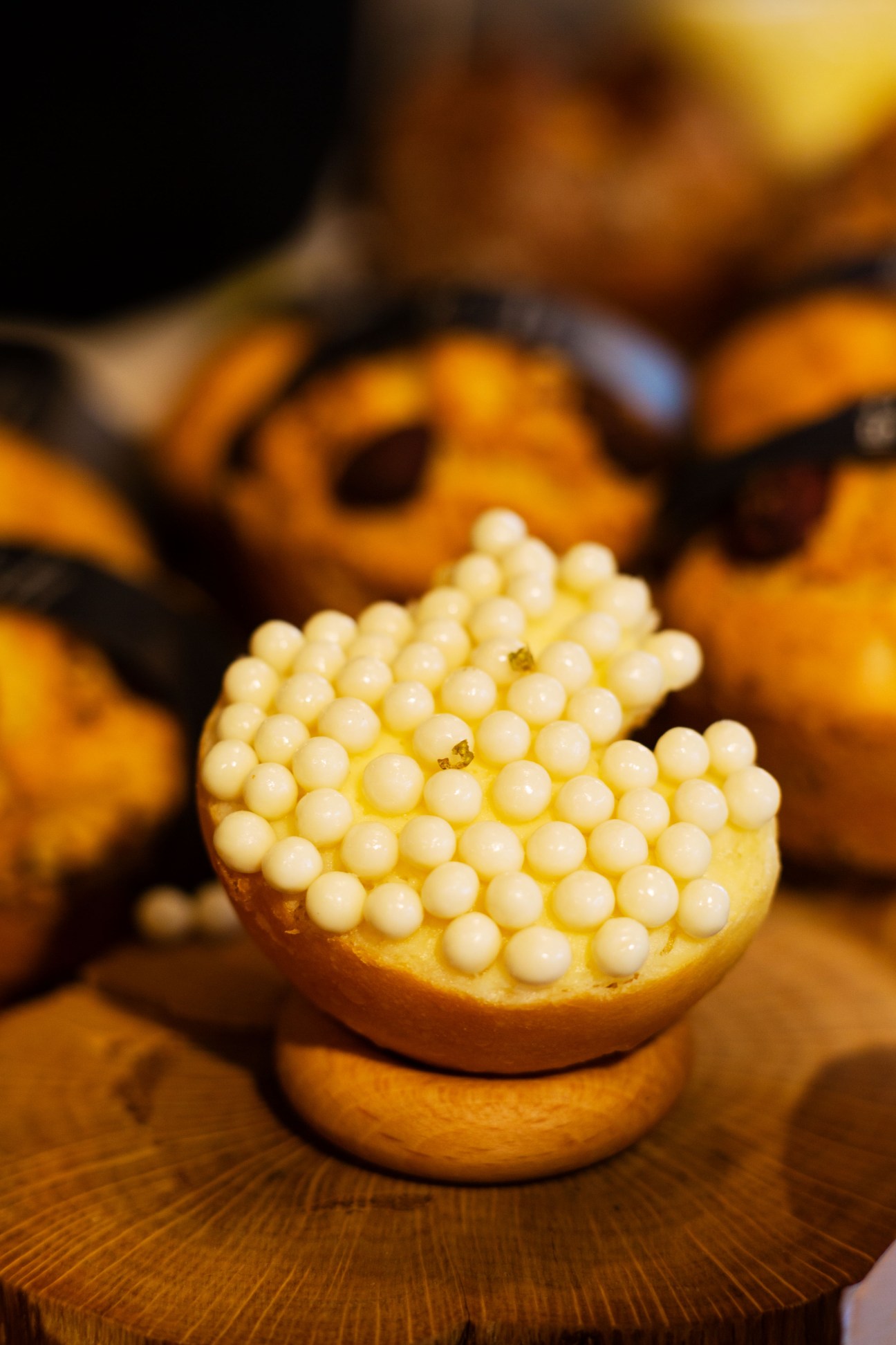 A pastry bun topped with white pearl-shaped cream beads on a wooden surface with blurred pastries in background.