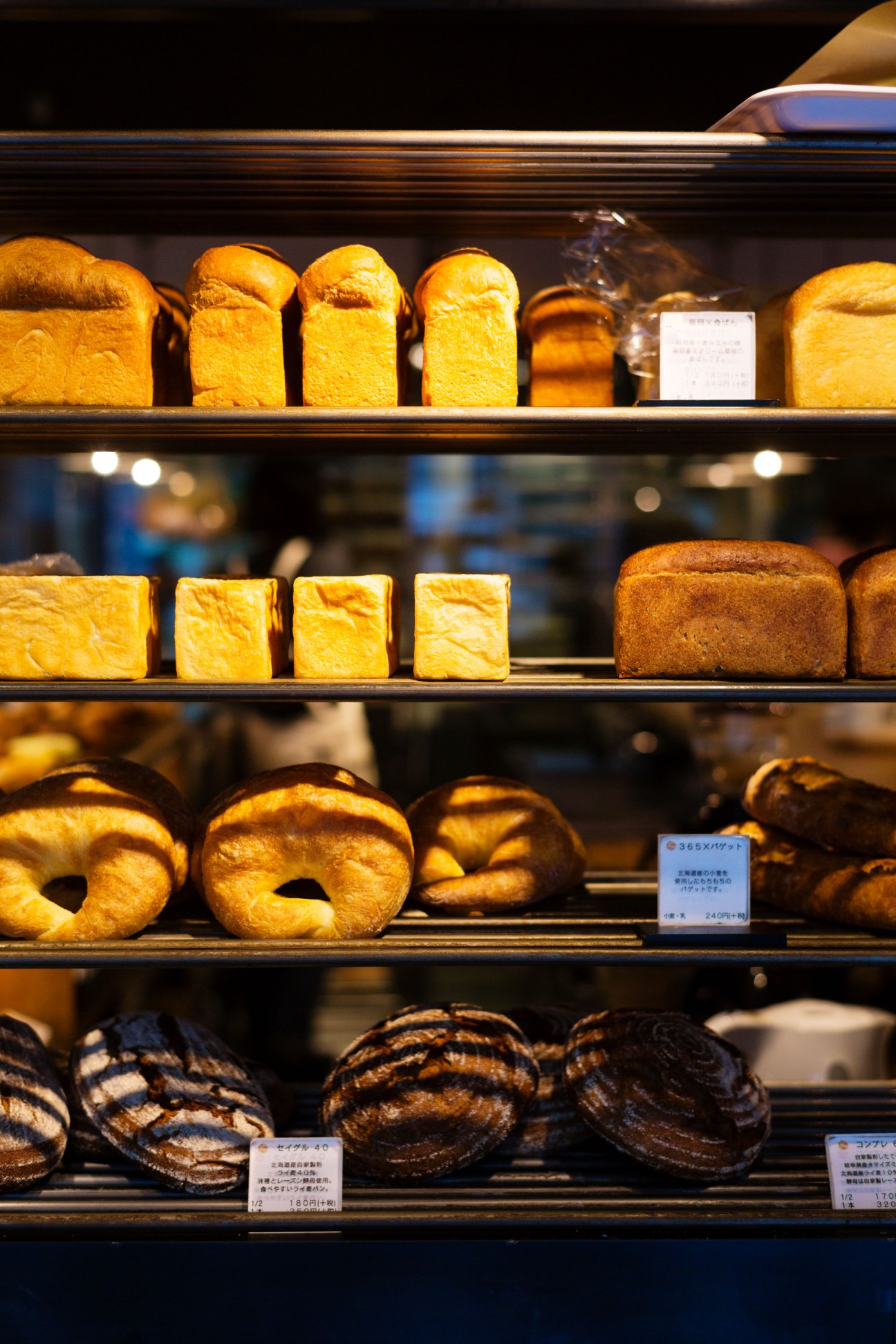 Illuminated bakery display shelves showcasing various golden-brown breads including loaves, rolls and bagels with Japanese price labels.