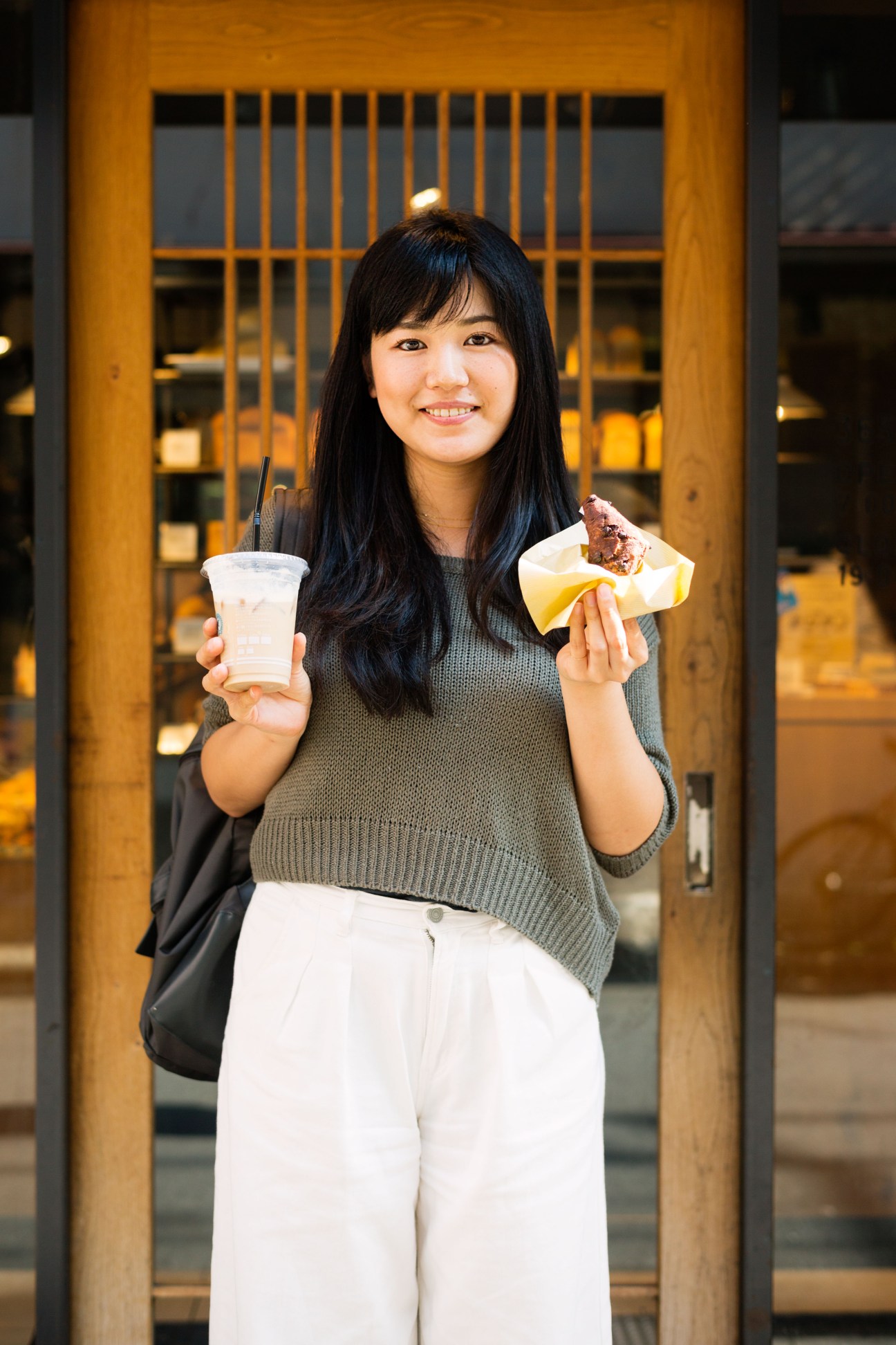 Young woman holding a beverage and chocolate bread pastry at a bakery storefront.