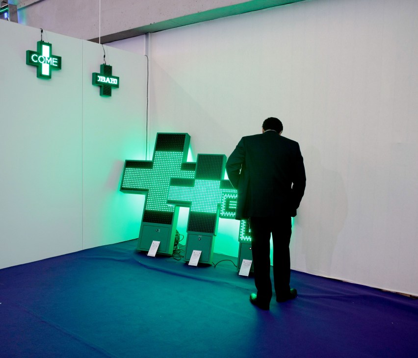 Man in dark suit viewing illuminated green pharmacy cross neon signs at trade fair.